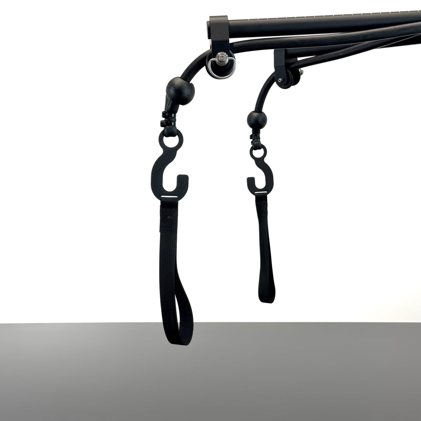 Black metal SlingShot camera mount arms ending with two rubber elastics knotted with black rubber stoppers and metal hooks with loops hanging at the end, shown against a white background above a grey table.